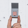 Eine Hand mit Nailart hält eine geöffnete graue Schachtel mit The Single Blush von LH Cosmetics vor einer weißen, strukturierten Wand.
