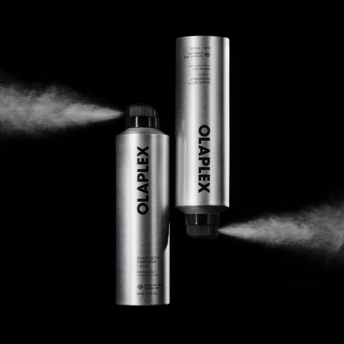 Zwei silberne OLAPLEX Shape Set Hairspray-Sprühdosen, aus denen Haarspray vor einem schwarzen Hintergrund versprüht wird.