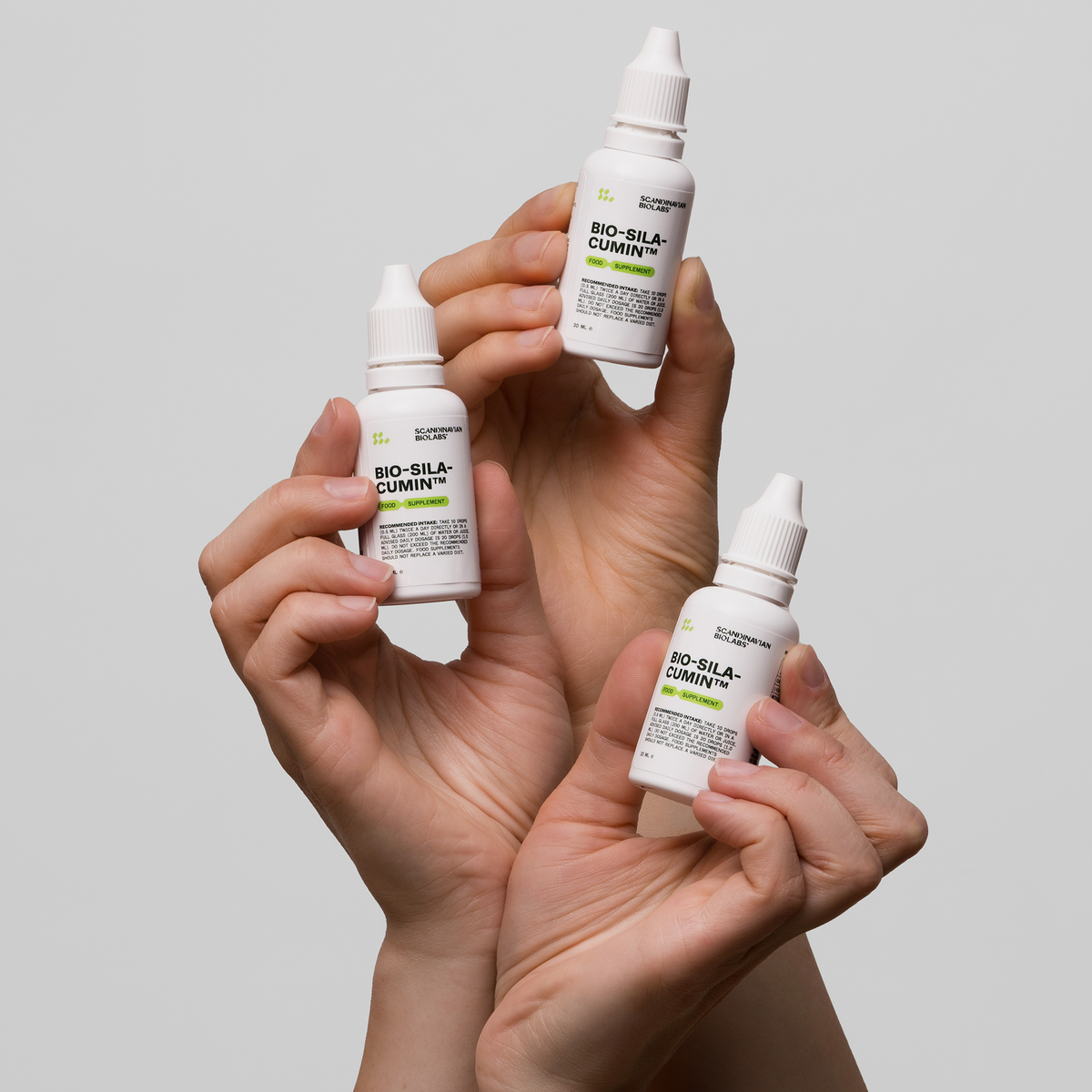 Drei Hände halten kleine weiße Flaschen mit der Aufschrift Bio-Silacumin von Scandinavian Biolabs vor einem schlichten hellgrauen Hintergrund.