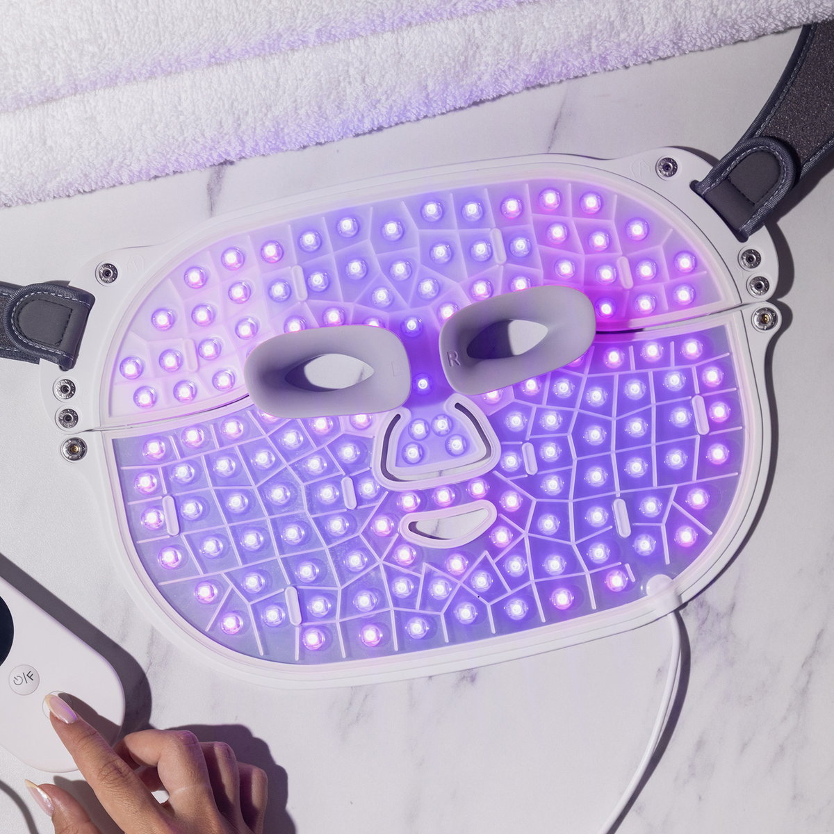 Die STYLPRO Wavelength Pro 5 in 1 LED-Maske leuchtet violett auf einer Marmoroberfläche, während eine Hand die angeschlossene Fernbedienung hält.