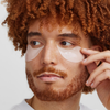 Eine Person mit lockigem Haar trägt XLASH Awakening Eye Gels auf und blickt dabei mit neutralem Gesichtsausdruck zur Seite.
