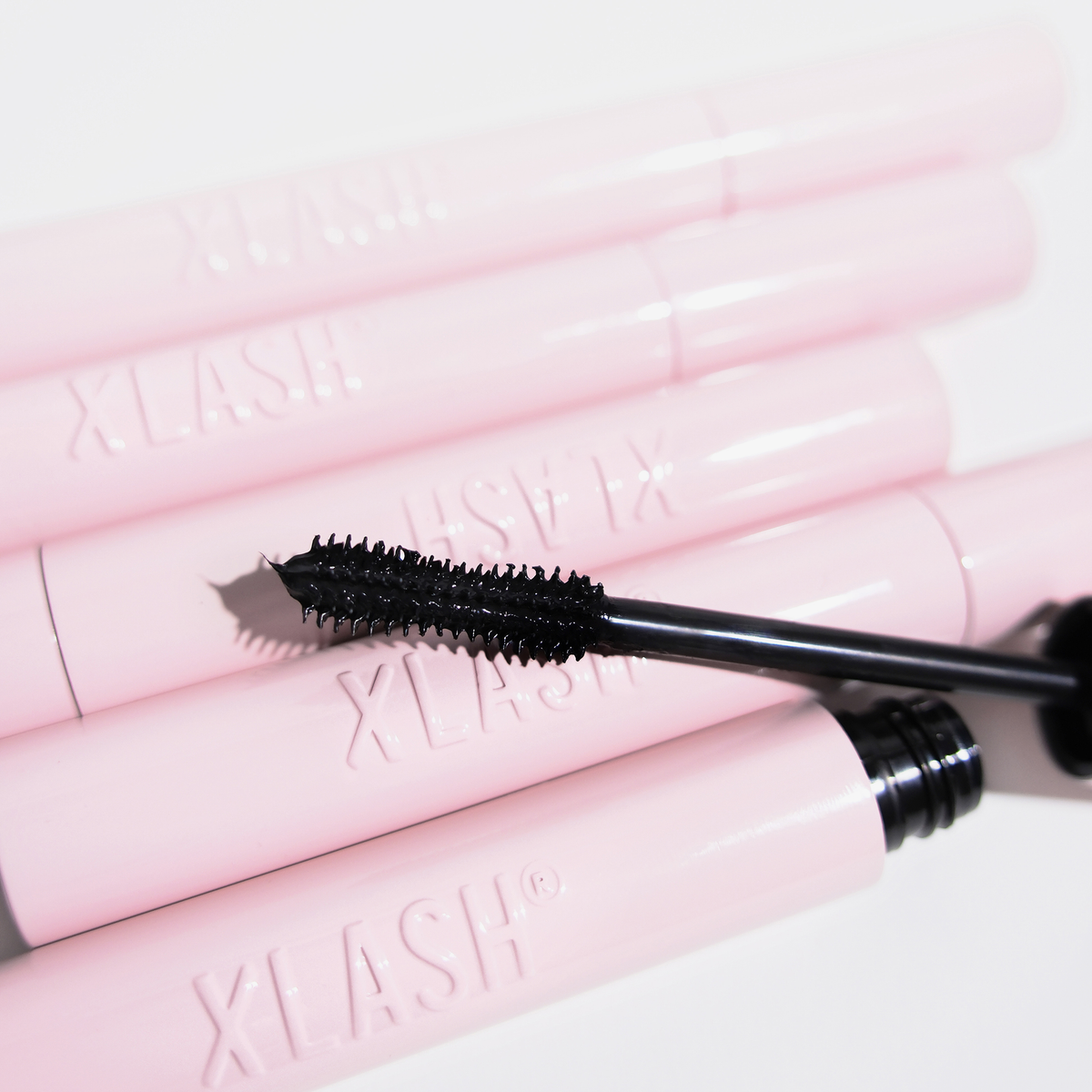 Auf weißem Hintergrund liegt eine schwarze Lash Hero-Mascara-Bürste auf rosa XLASH-Tuben.