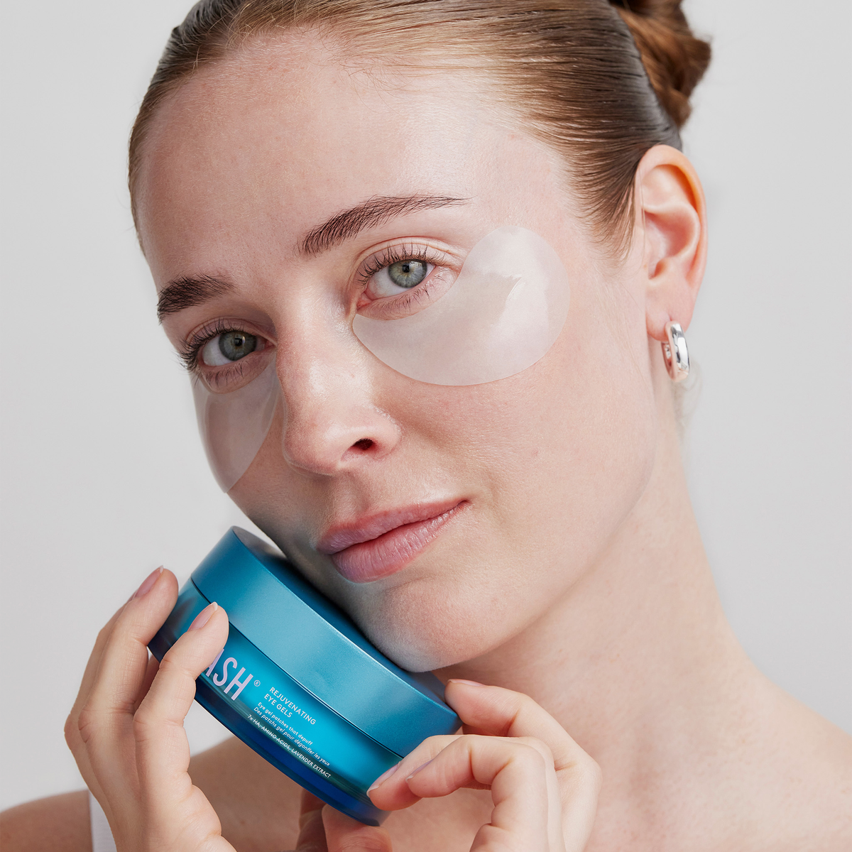 Eine Frau mit den Rejuvenating Eye Gels von XLASH hält ein blaues Töpfchen an ihre Wange und blickt in die Kamera.