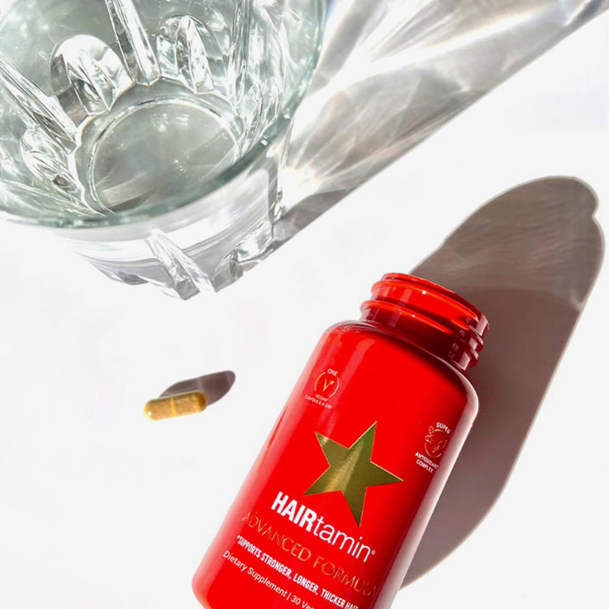 Eine rote Flasche PURISH Kevin Testproduct, eine gelbe Kapsel und ein Glas Wasser auf einer weißen Fläche mit Schatten.