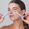 Eine Frau mit geflochtenen Haaren zwinkert, während sie das „BLONDESISTER 2 IN 1 EYES FIRST LASH&BROW SERUM“ vor einem schlichten Hintergrund nahe an ihre Lippen hält.