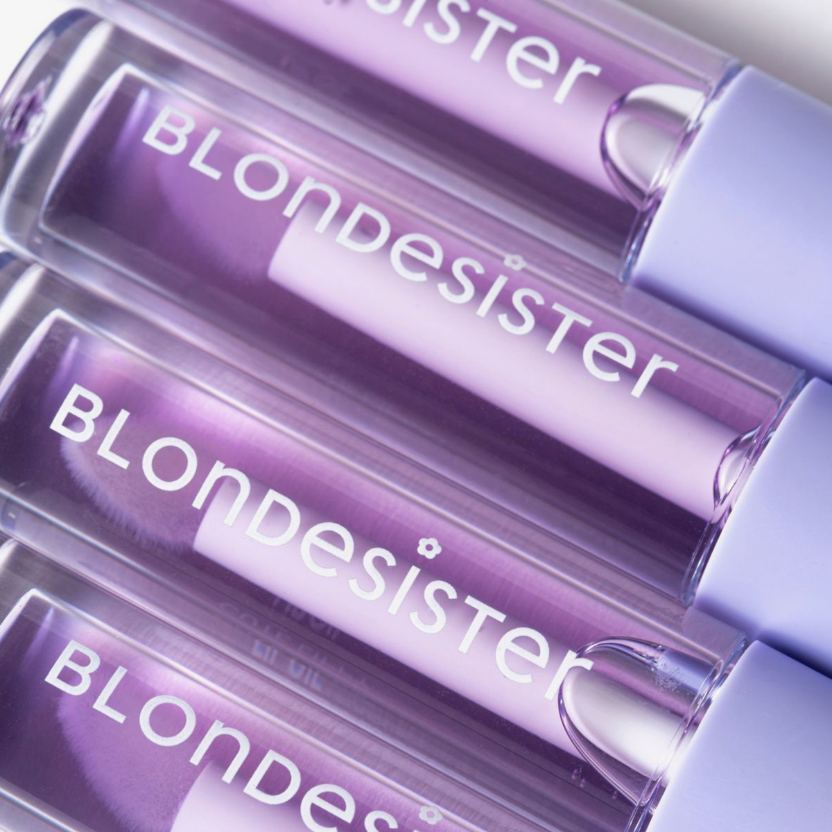 Vier Tuben „BLONDESISTER 3 IN 1 CUTE LIPPY LIP OIL“ in Lavendelfarbe, diagonal auf einem weißen Hintergrund angeordnet.