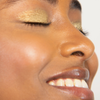 Eine Frau mit leuchtend goldenem GLOWEYE-LIDSCHATTEN von BLONDESISTER, glatter Haut und einem strahlenden Lächeln, die Augen vor Glückseligkeit geschlossen.