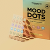 Ein Bogen mit bunten Mood Dots-Akne-Pflastern in Smiley-Form, ausgestellt vor der Verpackung ihres Produkts breakout+aid.