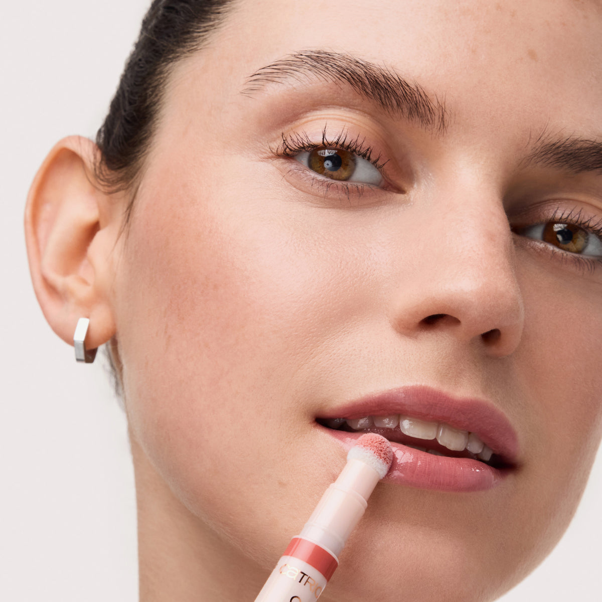 Eine Frau trägt Colour Cushion Juicy Lip Oil von Catrice Cosmetics auf ihre Lippen auf. Sie trägt natürliches Make-up und einen silbernen Creolenring.