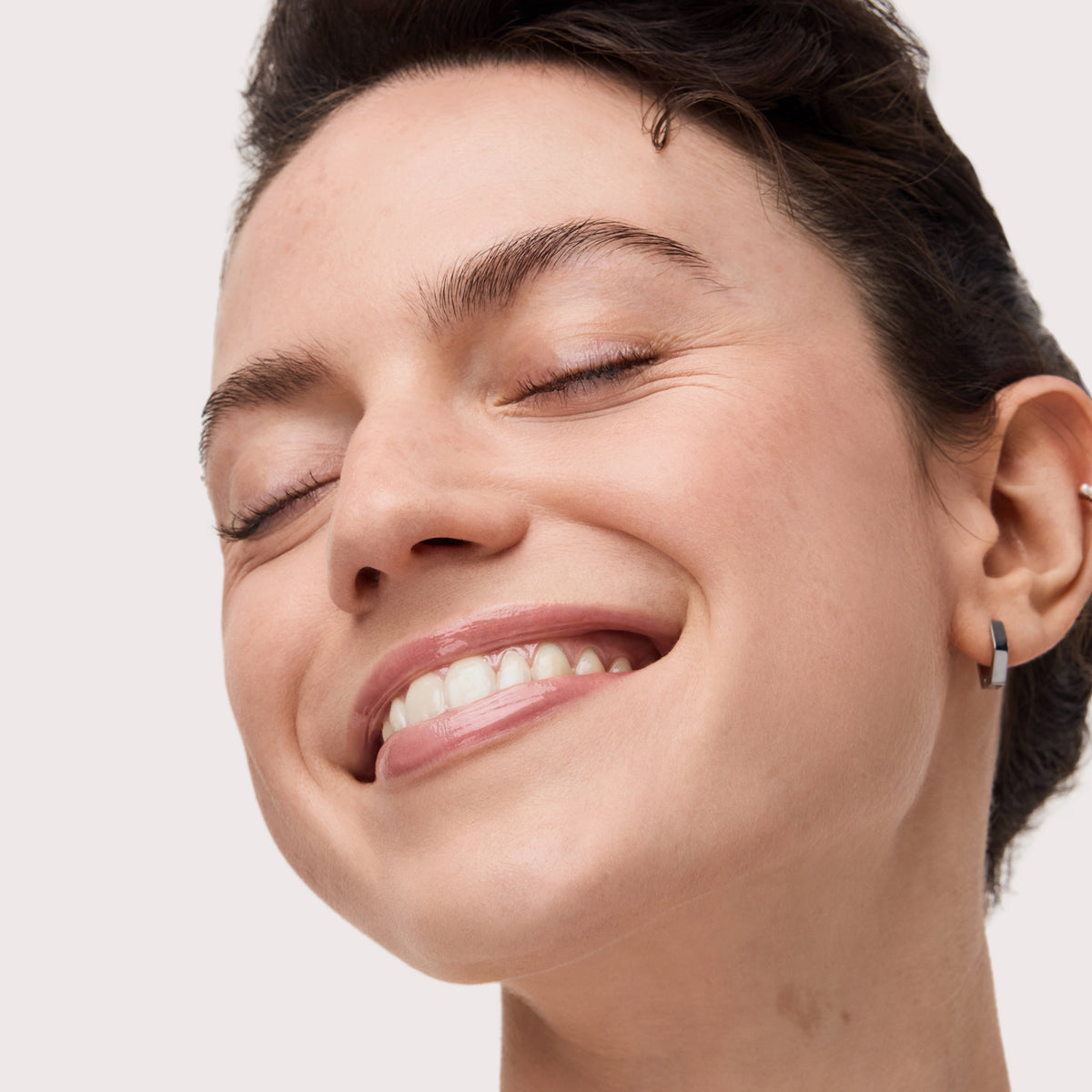 Eine Frau mit kurzen dunklen Haaren lächelt mit geschlossenen Augen, sie trägt einen kleinen Ohrring und Colour Cushion Juicy Lip Oil von Catrice Cosmetics.
