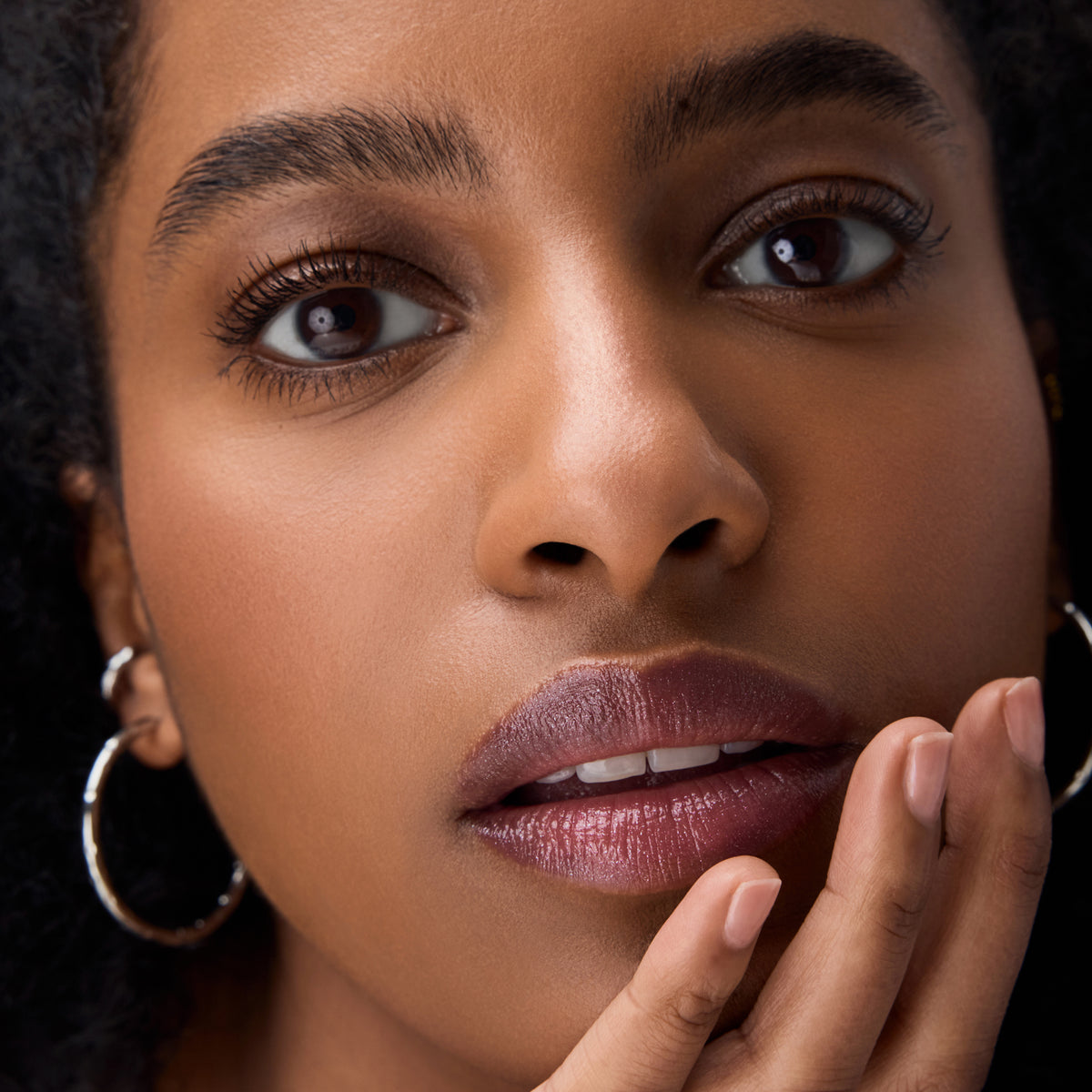 Nahaufnahme eines Gesichts mit schimmernden Lippen in Beerenrosa, natürlichem Augen-Make-up und silbernen Creolen, eine Hand berührt sanft das Kinn.
