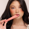 Frau mit langen dunklen Haaren hält einen rosafarbenen Juicy Lip Balm von Dominique Cosmetics an ihre Lippen und schaut in die Kamera.