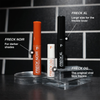 Der Freck OG Sommersprossenstift von Freck Beauty wird aufrecht auf einem durchsichtigen Ständer vor einem schwarzen Kachelhintergrund gezeigt.