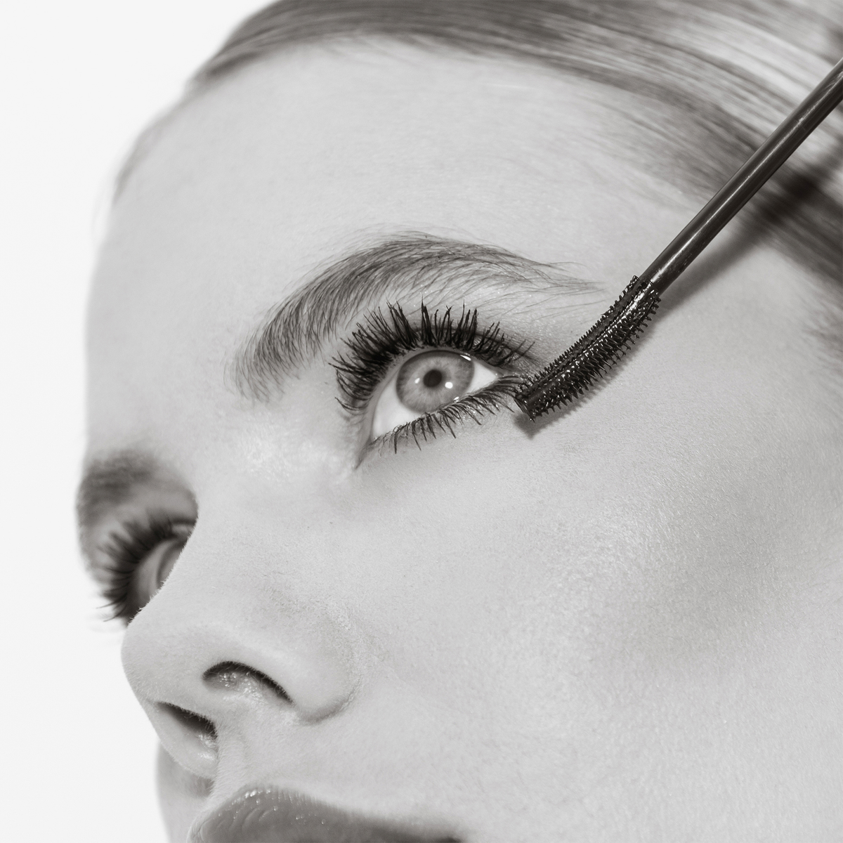 Schwarz-Weiß-Nahaufnahme einer Frau, die THE GLAM FEMULINE Mascara von Mesauda Beauty mit einem Zauberstab auf ihre Wimpern aufträgt.