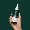 Vor einem dunkelgrünen Hintergrund hält eine Hand die „Philip Kingsley Density Preserving Scalp Drops“ in der Hand.