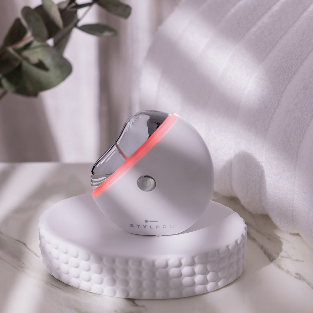 Weißer STYLPRO Gua Sha mit silberner Fläche und rotem Licht, elegant platziert auf einem Podest im Badezimmer-Ambiente.