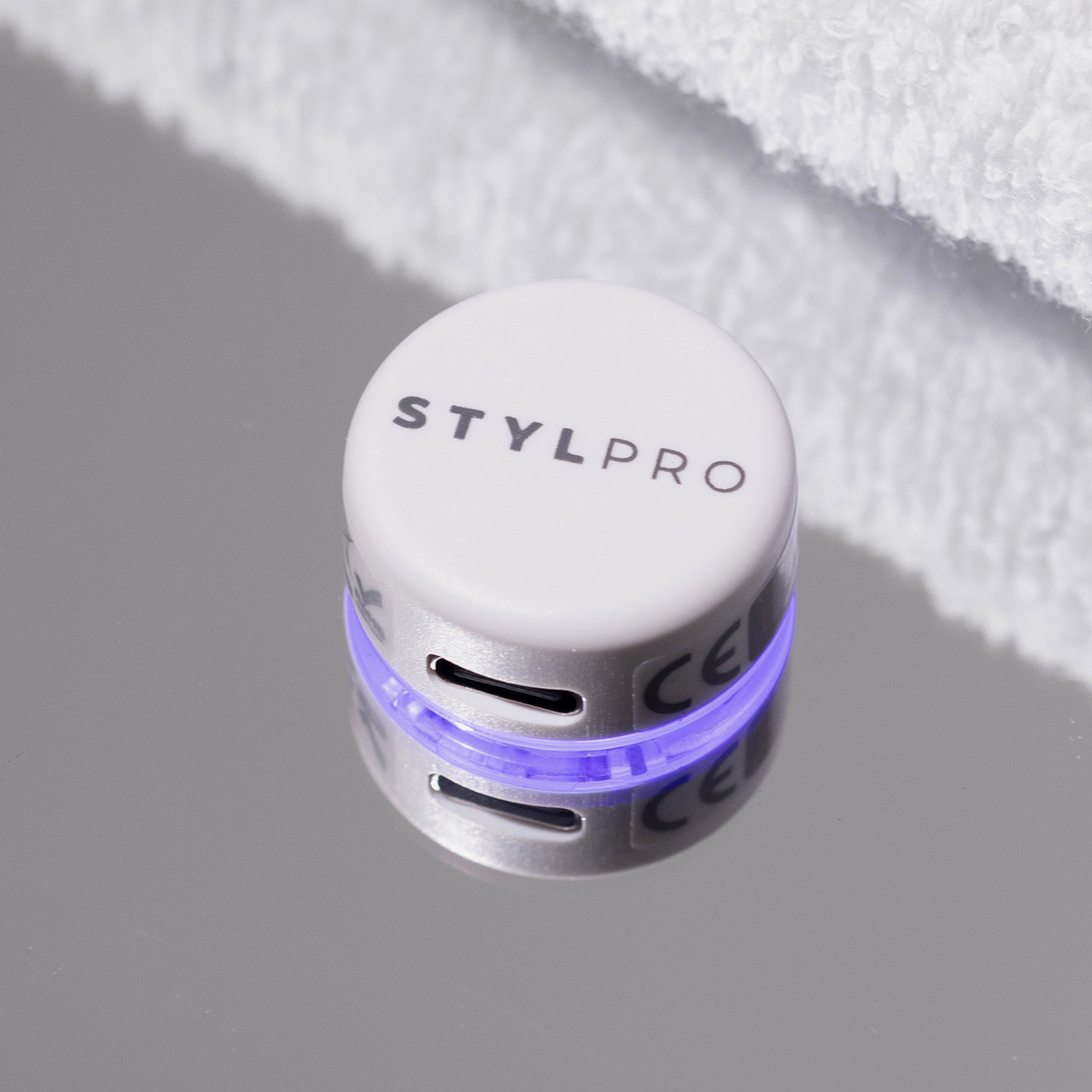 Das STYLPRO LED Spot & Blemish Patch, ein weißes, rundes Gerät mit blauem Licht, wird auf einer reflektierenden Oberfläche neben einem weißen Handtuch angebracht.