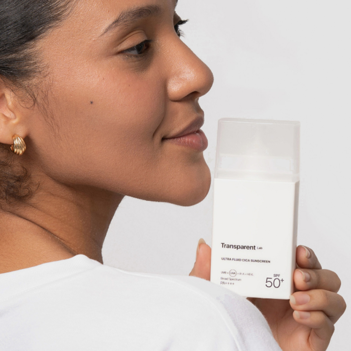 Eine lächelnde Frau, die eine weiße Flasche „Transparent Lab PHA Soft Peeling Cleanser Mini“ vor ihrem Gesicht hält, vor einem hellen Hintergrund.