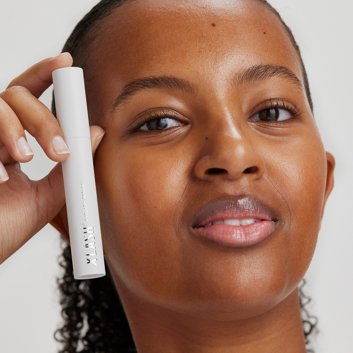 Eine Frau lächelt und hält eine Tube Xlash Eyelash Serum vor ihr Gesicht, vor einem schlichten Hintergrund.
