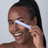 Eine lächelnde Person hält eine Tube „BLONDESISTER 2 IN 1 EYES FIRST LASH&BROW SERUM“ vor ihrem Auge vor einem schlichten, hellen Hintergrund.