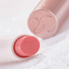 Nahaufnahme des Catrice Diamond Glaze Gloss Stick in einem schimmernden Korallton, liegend mit sichtbarer Produktspitze und rosigem Deckel im Hintergrund.