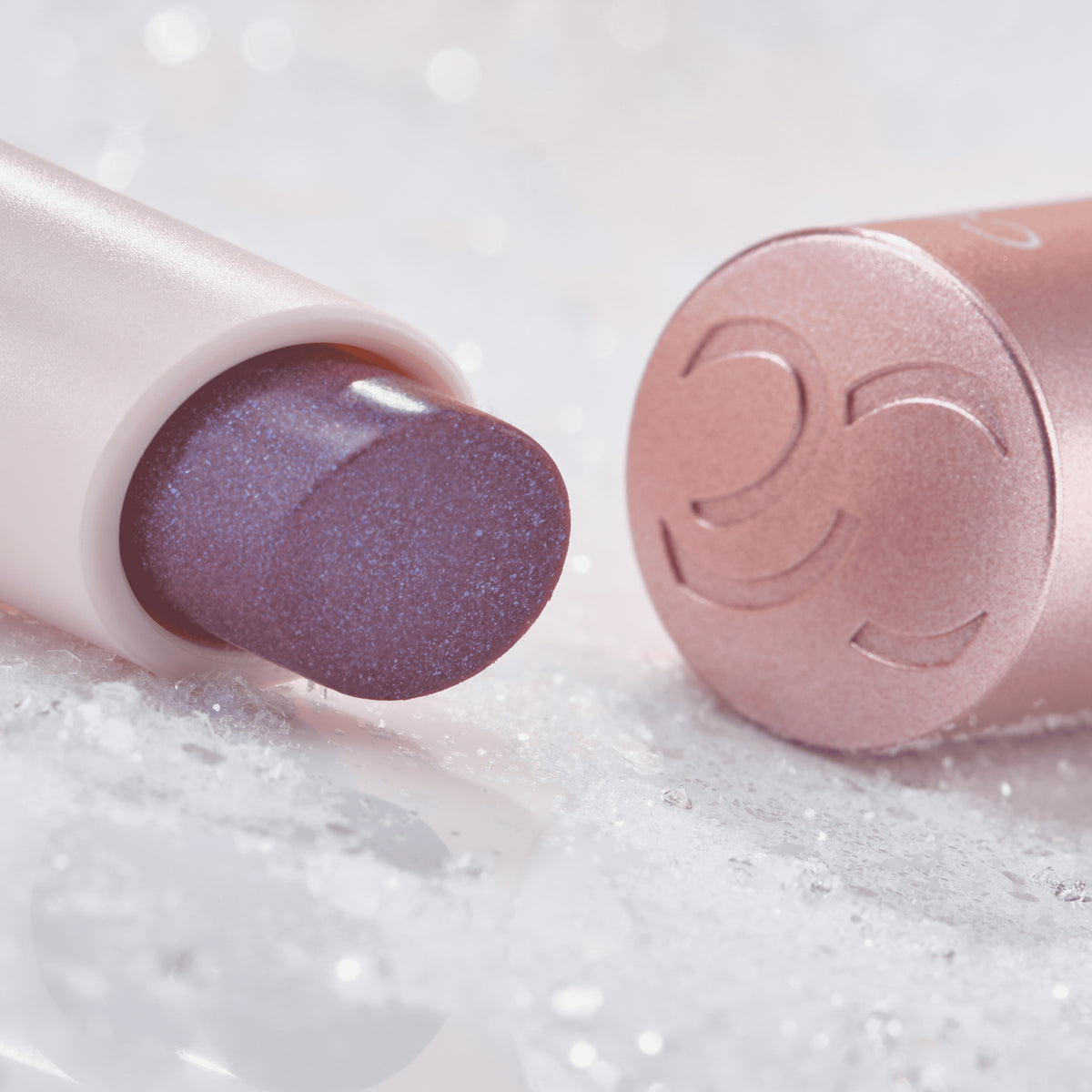 Nahaufnahme des Catrice Diamond Glaze Gloss Stick in einem schimmernden Lilaton, liegend mit sichtbarer Produktspitze und rosigem Deckel im Hintergrund.