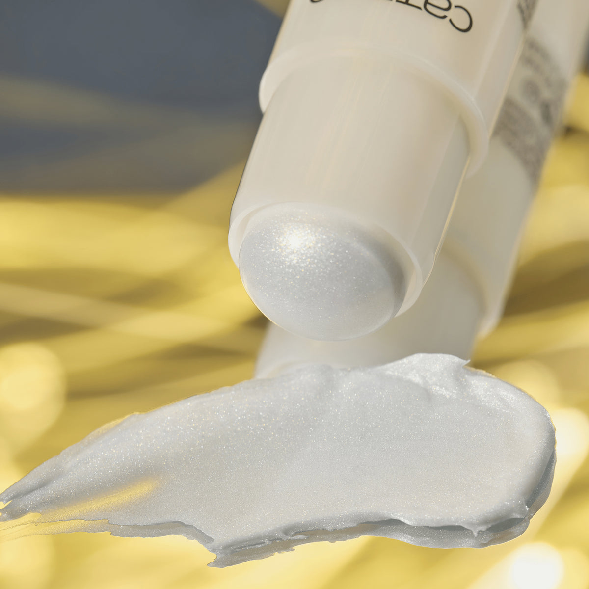 Nahaufnahme eines schimmernden Highlighter-Sticks von Catrice mit aufgetragener cremiger Textur in Perlweiß vor goldenem Hintergrund.