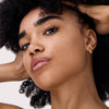 Eine Frau mit lockigem Haar, Sommersprossen, goldenen Ohrringen und natürlichem Make-up posiert mit den Händen im Haar.