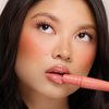 Frau mit glänzenden Lippen und pfirsichfarbenem Rouge hält Dominique Cosmetics Juicy Lip Balm in der Nähe ihres Mundes und blickt nach oben.