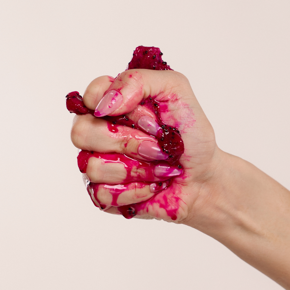 Eine Hand mit rosafarbenen Nägeln drückt den Juicy Lip Balm von Dominique Cosmetics, roter Glanz tropft an den Fingern herunter.