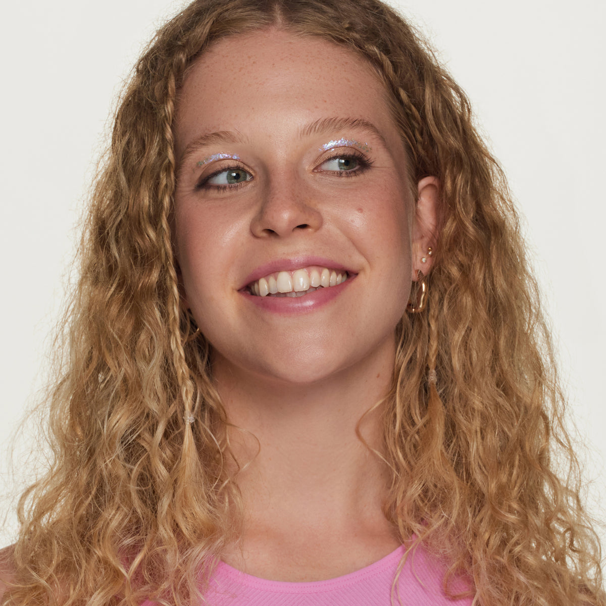 Eine junge Frau mit lockigem, blondem Haar lächelt seitlich. Sie trägt schimmernden Lidschatten, dezenten Lipgloss und ein pinkes Oberteil.
