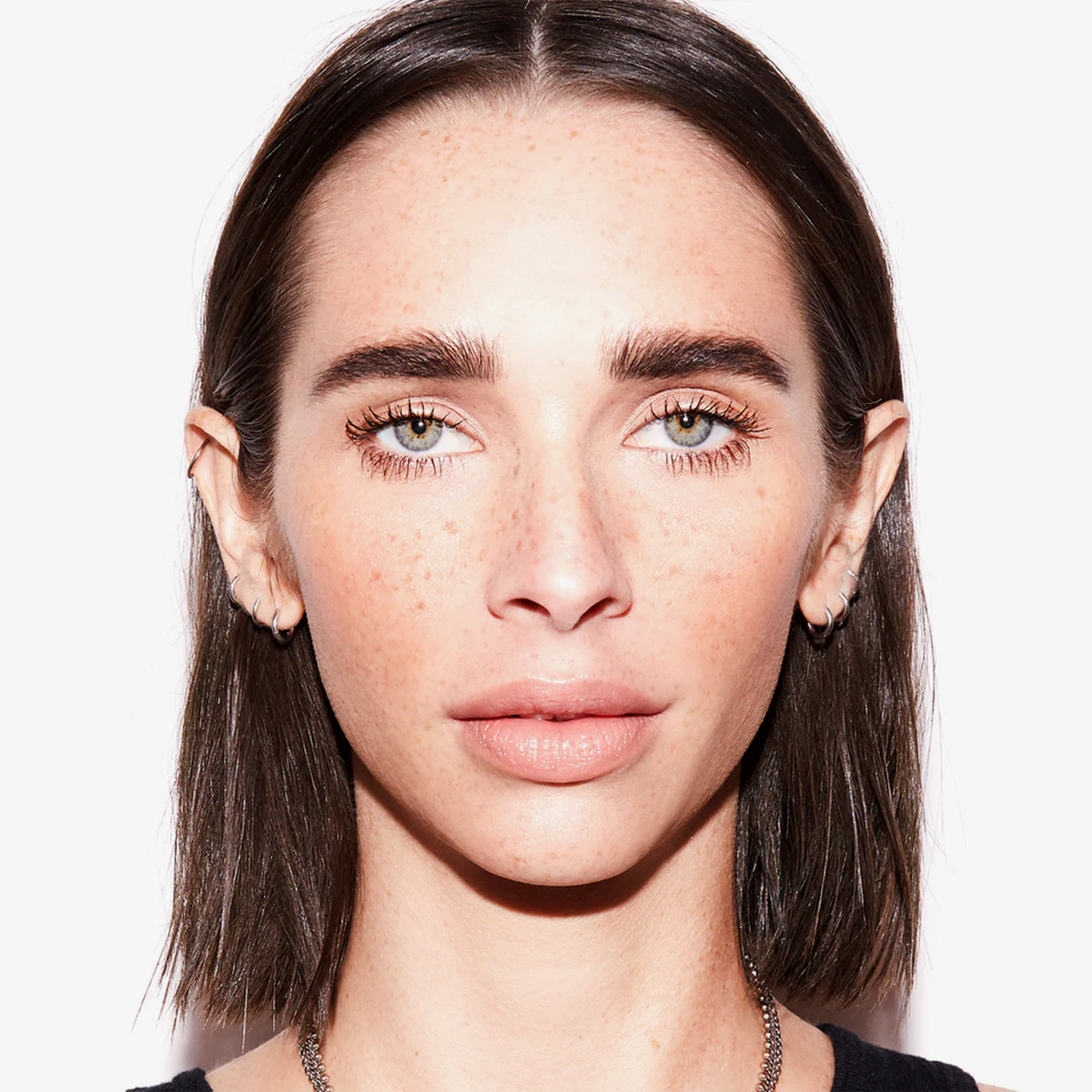 Eine Person mit glattem dunkelbraunem Haar und Sommersprossen trägt Freck OG von Freck Beauty und blickt nach vorne vor einem weißen Hintergrund.