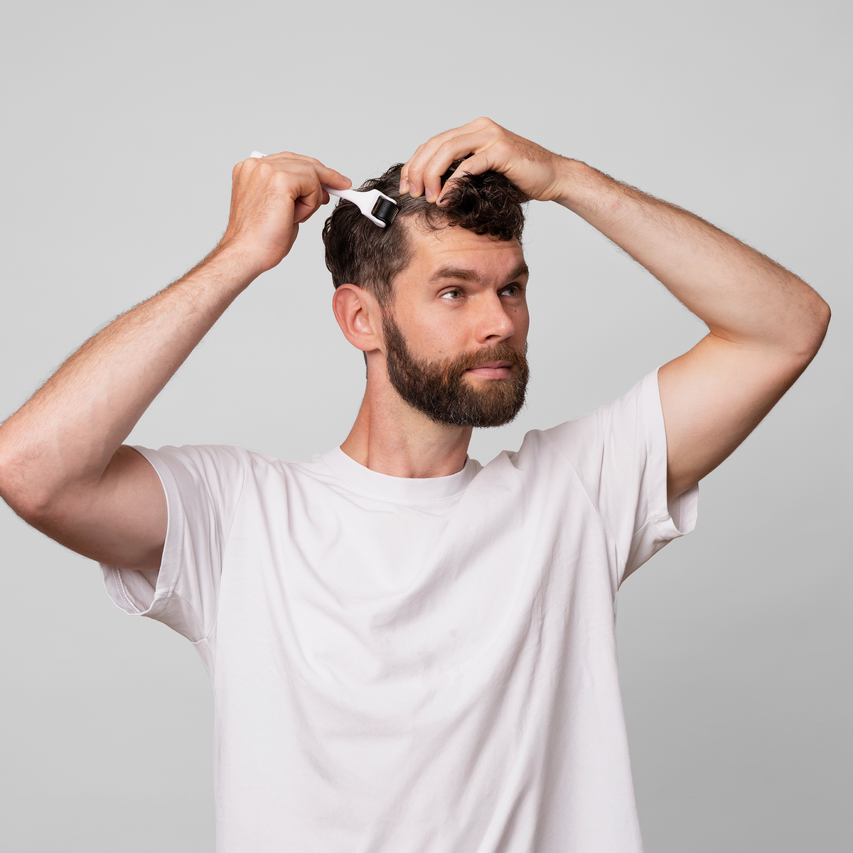 Ein bärtiger Mann in einem weißen T-Shirt benutzt den Scandinavian Biolabs Scalp Activation Derma Roller und bürstet sein Haar vor einem schlichten hellgrauen Hintergrund.