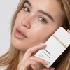 Eine Frau mit reiner Haut hält die getönte Sonnencreme „Transparent Lab Mineral Tinted Sunscreen“ dicht an ihr Gesicht und blickt in die Kamera.