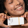 Eine lächelnde Person hält eine weiße, getönte Sonnencreme von Transparent Lab mit der Aufschrift „SPF 50“ dicht an ihr Gesicht.