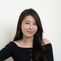 Junge asiatische Frau mit dunklem gewelltem Haar und Sommersprossen, die ein schwarzes schulterfreies Oberteil trägt, lächelt leicht in die Kamera.