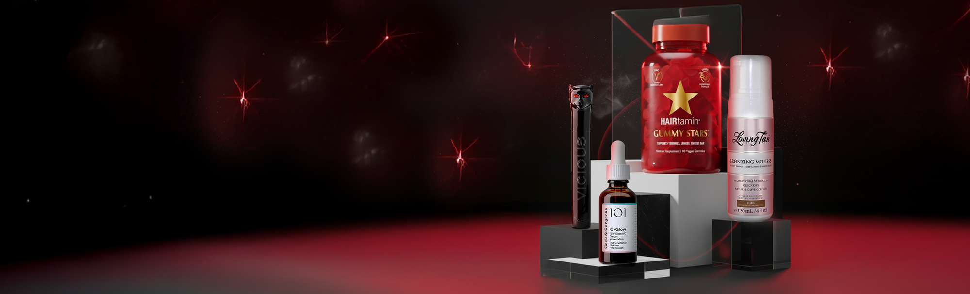 Vier Beauty-Produkte auf schwarzen Podesten vor dunklem Hintergrund mit roten Lichtakzenten: Geek & Gorgeous C-Glow Serum, Hairtamin Gummy Stars, NABLA Vicious Mascara und Loving Tan Bronzing Mousse.