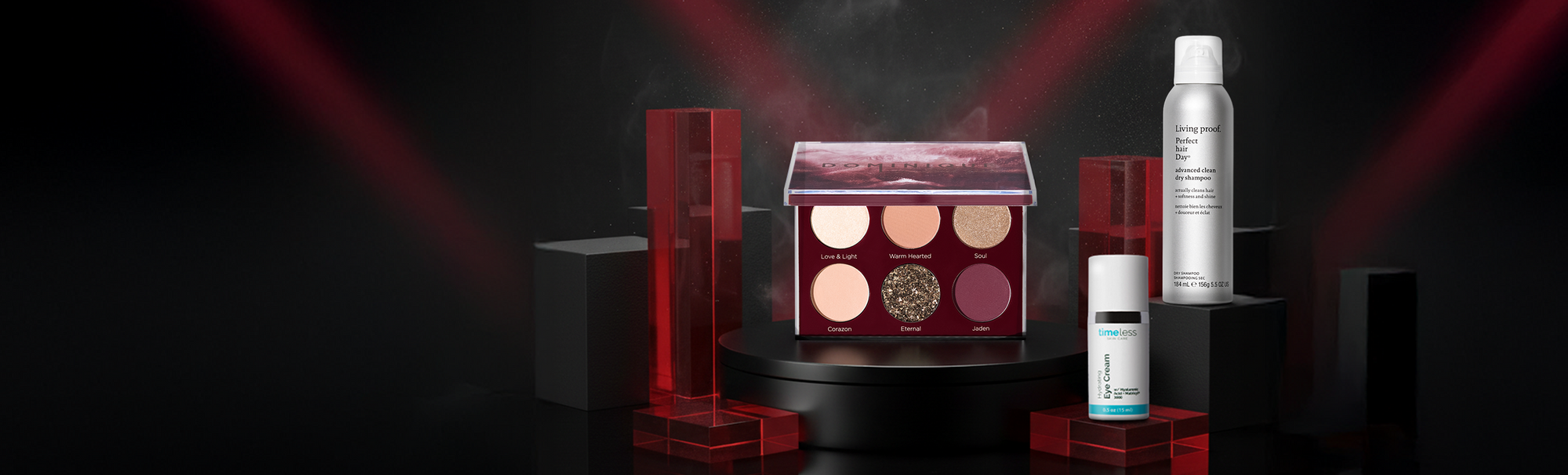 Drei Beautyprodukte auf schwarzen Podesten vor dunklem Hintergrund mit Lichteffekten: Dominique Cosmetics Unconditional Palette, Timeless Skin Care Hydrating Eye Cream und Living Proof. Perfect Hair Day Advanced Clean Dry Shampoo.