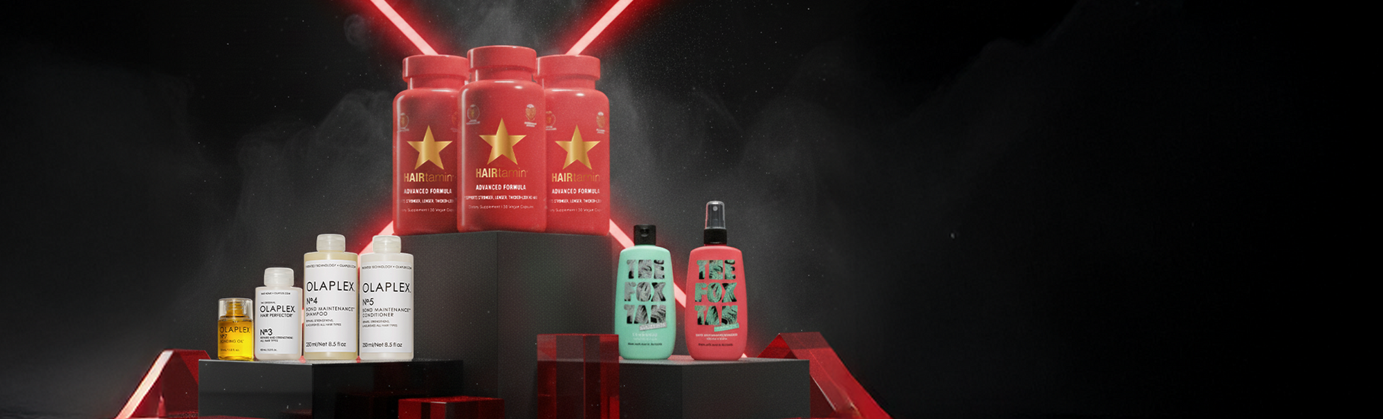 Drei Beauty Bundle auf schwarzen Podesten vor dunklem Hintergrund mit roten Lichtlinien: HAIRtamin Advanced Formula 3 Month Bundle, OLAPLEX Love Your Hair Bundle und The Best of The Fox Tan Bundle.