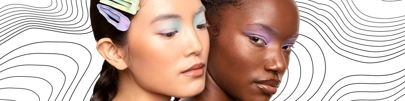 Zwei Frauen mit pastellfarbenem Lidschatten und Haarspangen posieren zusammen vor einem gewellten schwarz-weißen Hintergrund.