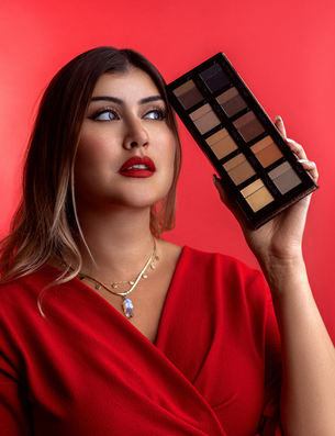 Eine Frau in rotem Kleid hält die Danessa Myricks Beauty Groundwork Palette mit neutralen Brauntönen vor einem roten Hintergrund.