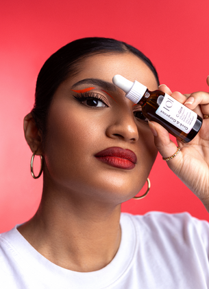 Eine Frau mit rotem Lippenstift und grafischem Eyeliner hält eine Flasche Geek & Gorgeous C-Glow Serum vor ihrem Auge.