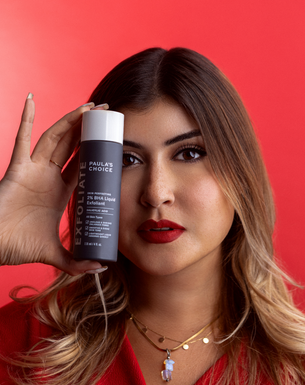 Eine Frau mit rotem Lippenstift hält eine Flasche Paula’s Choice 2% BHA Liquid Exfoliant neben ihr Gesicht vor rotem Hintergrund.