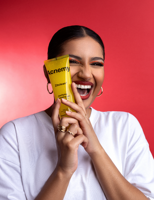 Eine lachende Frau in weißem Shirt hält ein gelbes Acnemy Zitclean Reinigungsgel vor ihr Gesicht vor rotem Hintergrund.