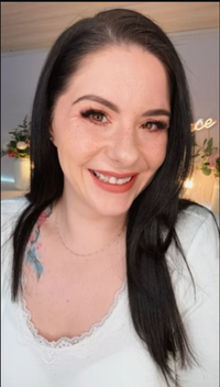 Frau mit langem dunklem Haar lächelt in die Kamera; heller Hintergrund mit Deko, Make-up betont Augen und Lippen, florales Tattoo am Schlüsselbein sichtbar.