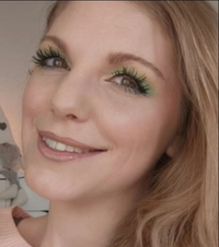 Frau mit blondem Haar lächelt in die Kamera; auffälliges grünes Augen-Make-up und dezente Lippenfarbe, weicher Hintergrund im Innenraum.