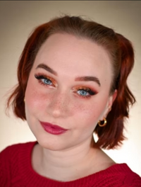 Frau mit kurzem rötlichem Haar und gold-rotem Augen-Make-up blickt in die Kamera; deutliche Sommersprossen, rosa Lippenfarbe, neutraler Hintergrund.