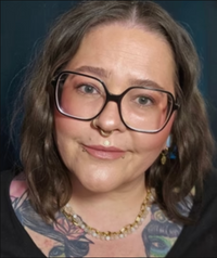 Frau mit dunklem, gewelltem Haar, großer Brille und dezentem Make-up blickt in die Kamera; sichtbare Tattoos und goldener Schmuck.