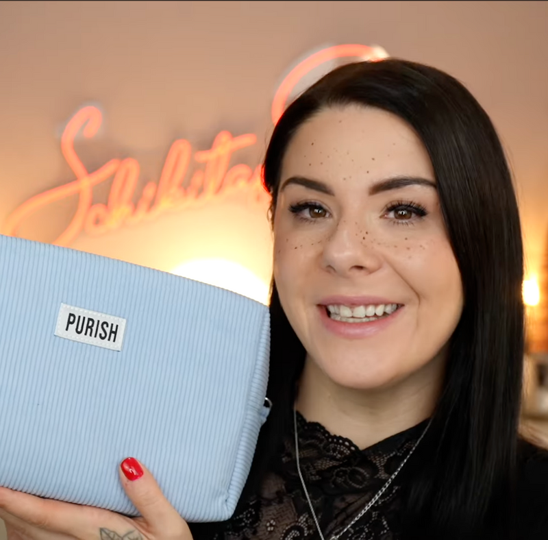 Frau mit dunklem Haar hält eine hellblaue Purish-Tasche in die Kamera und lächelt; unscharfer, warm beleuchteter Hintergrund.