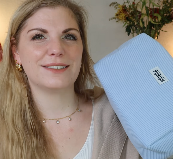 Frau mit blondem Haar hält eine hellblaue Purish-Tasche in die Kamera und lächelt; dezenter, heller Hintergrund mit Blumen.
